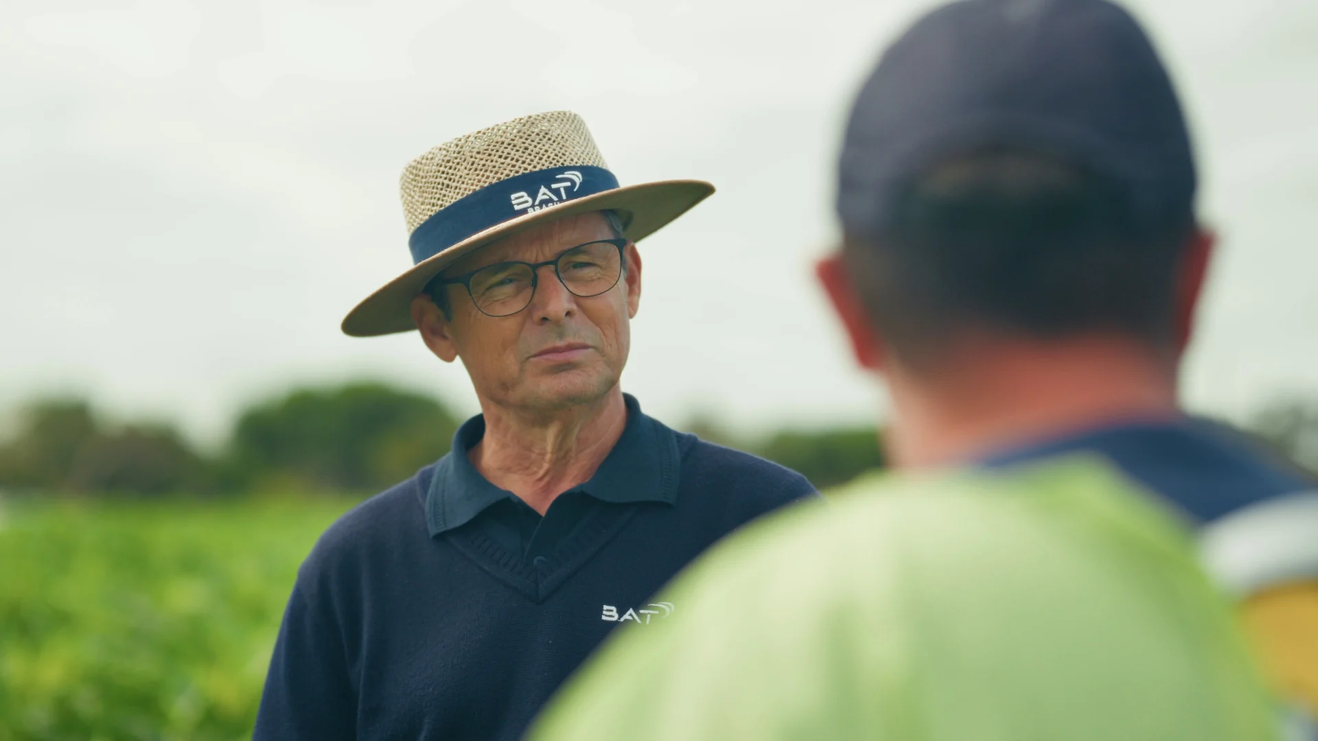 Orientador agrícola de chapéu de palha com a marca BAT conversa com um produtor na lavoura.