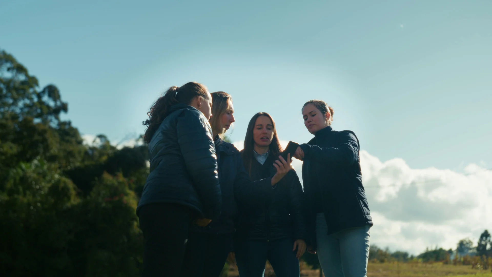 Quatro mulheres conversam ao ar livre, vistas de baixo contra o céu azul, com árvores ao fundo.