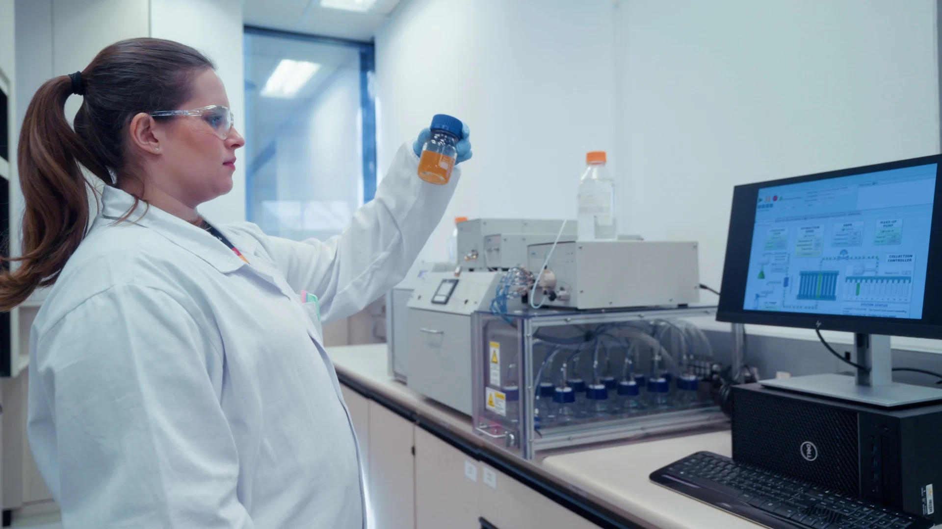 Pesquisadora de jaleco e óculos de proteção ergue um frasco com amostra ao lado de equipamentos e monitor em laboratório.