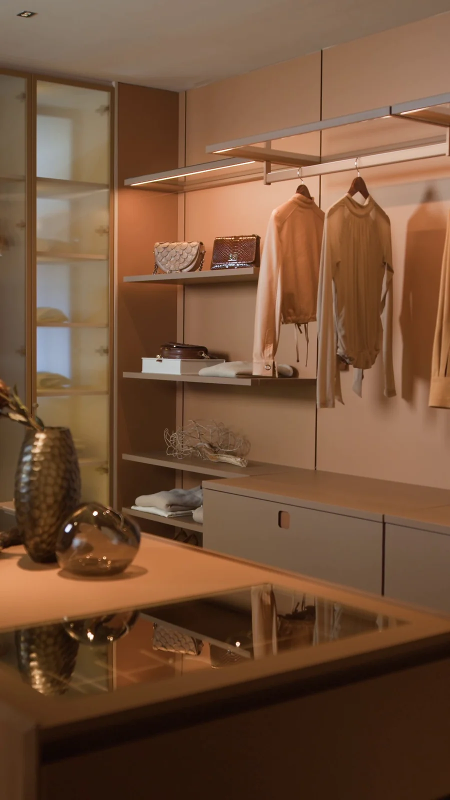 Closet em tons caramelo com roupas penduradas, prateleiras iluminadas, vitrine de vidro e ilha central com tampo espelhado.