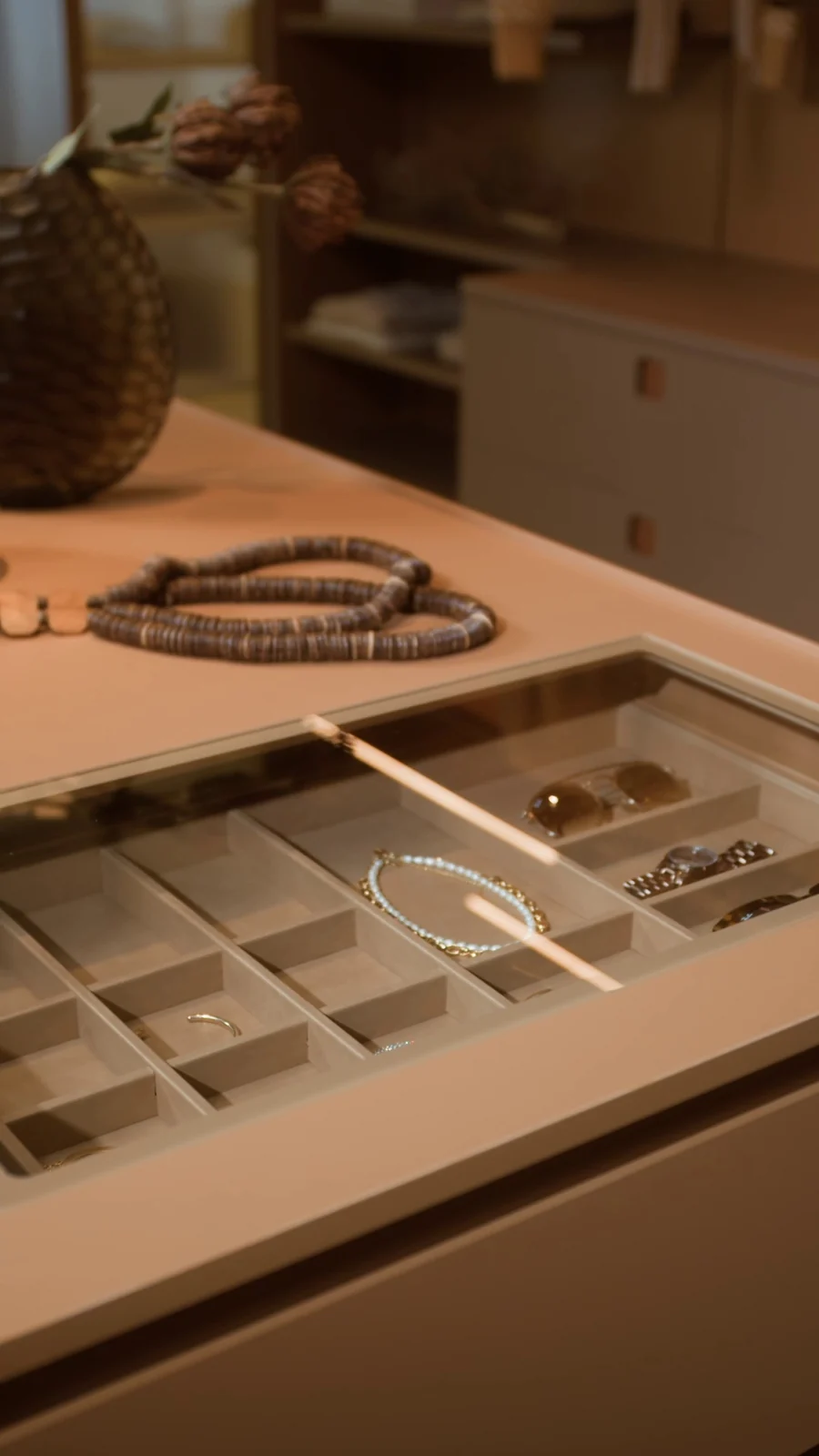 Gaveta de joias aberta na ilha do closet, com divisórias forradas, pulseira e óculos, sob um colar de contas sobre o tampo.