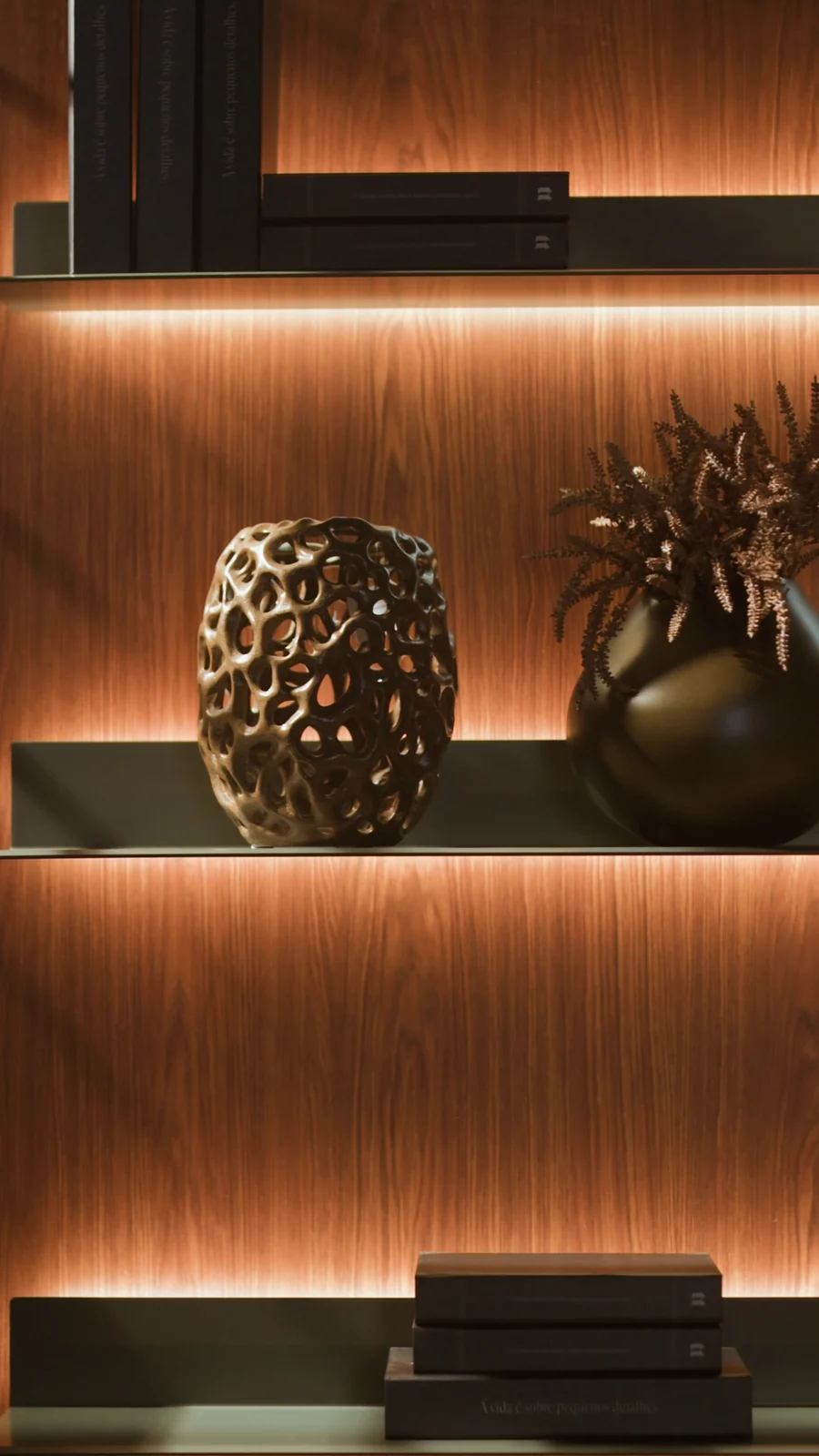 Prateleiras em madeira escura com iluminação embutida exibem vaso vazado dourado, arranjo seco e livros.
