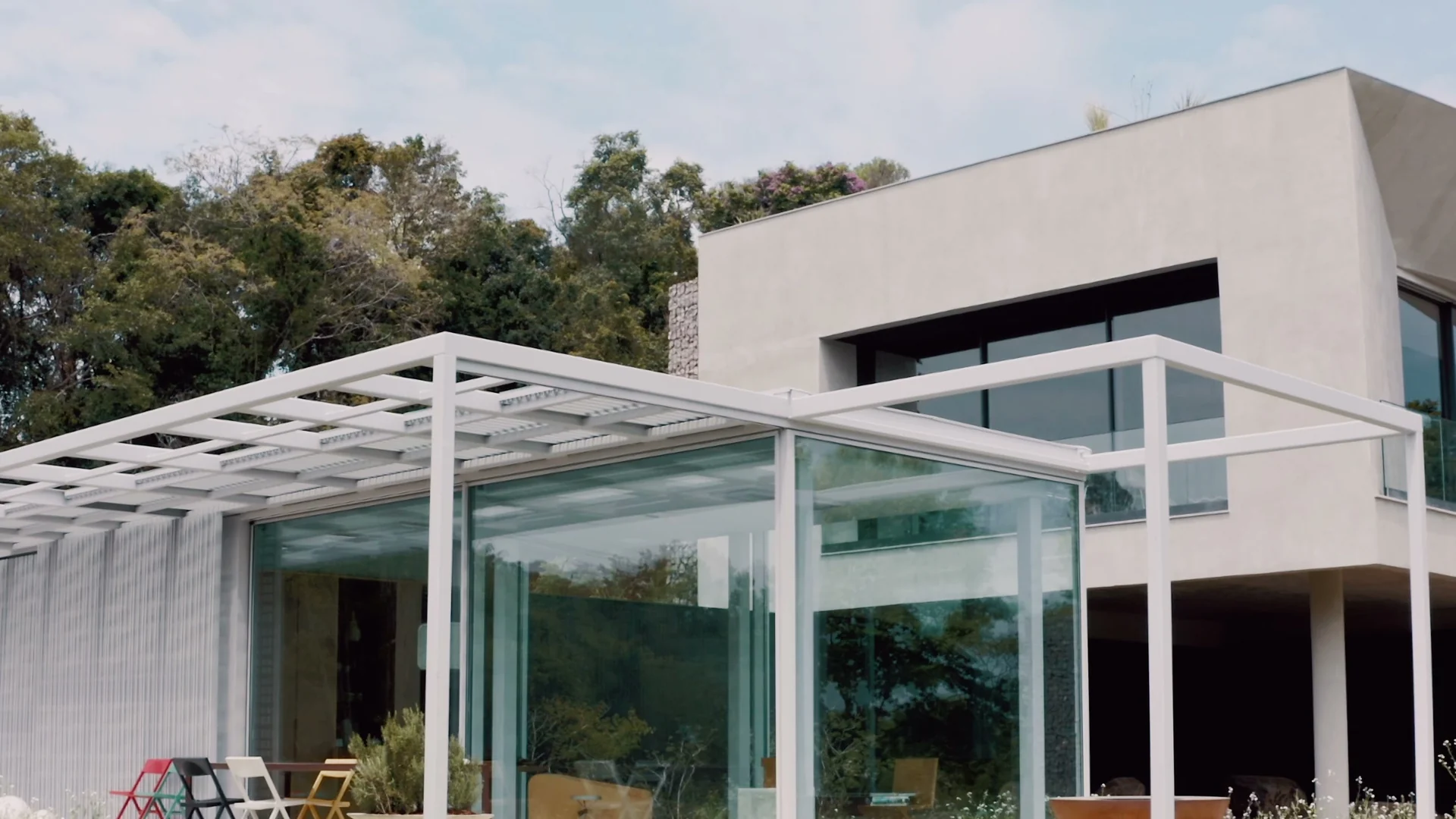 Casa de concreto com pavilhão de vidro e pérgola branca, vista de frente e cercada de mata.
