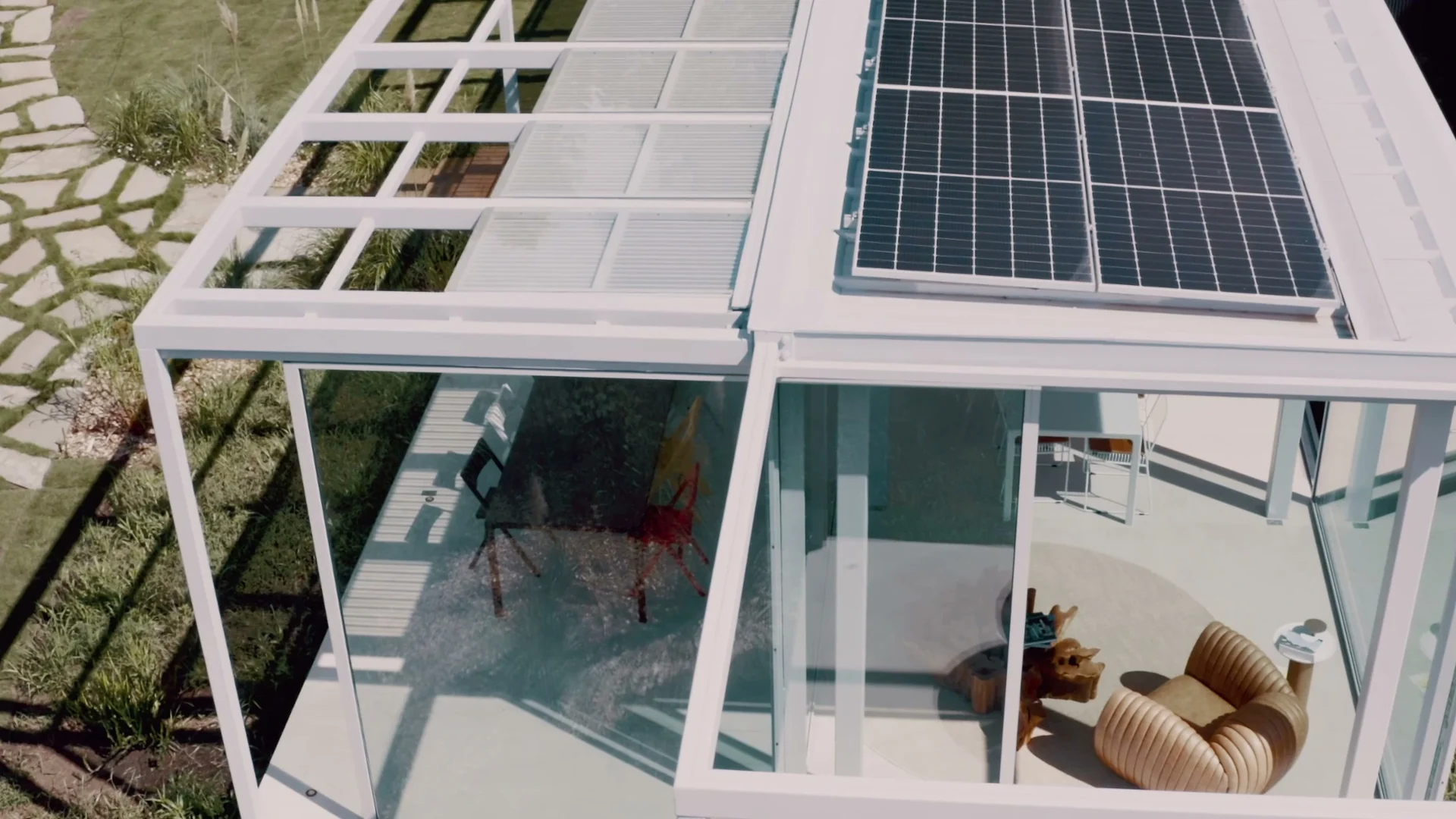Vista aérea do pavilhão de vidro com painel solar no teto e uma poltrona de madeira ondulada visível por dentro.