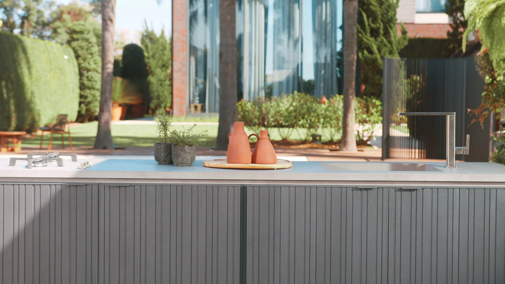 Frente da bancada ranhurada com vasos de terracota sobre uma bandeja de madeira e um jardim com palmeiras desfocado ao fundo.
