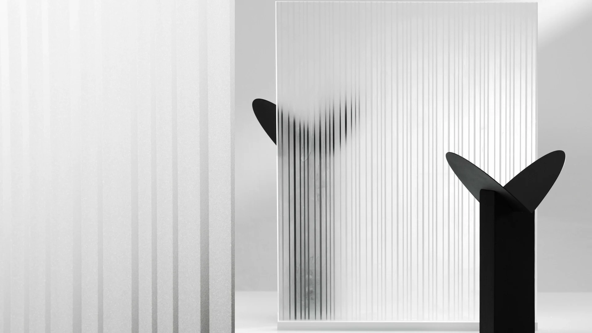 Painel de vidro com estampa de listras verticais diante de uma escultura preta em forma de duas folhas, em fundo branco.