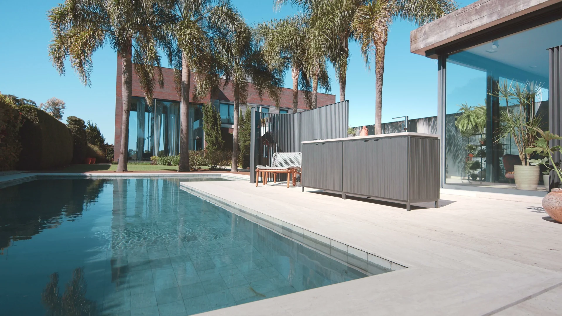 Piscina de borda reta sob sol forte, com palmeiras ao fundo e uma bancada de cozinha externa em alumínio cinza ao lado de uma casa de vidro.