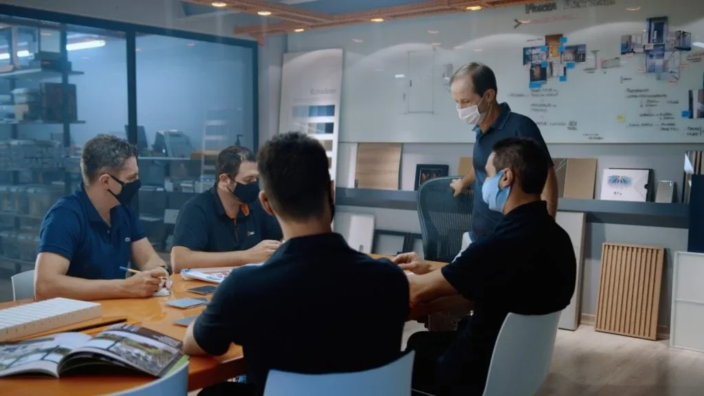 Equipe de projeto de máscara reunida em torno de uma mesa com amostras de materiais e catálogos, em cena do filme Veritas, da Cinex.