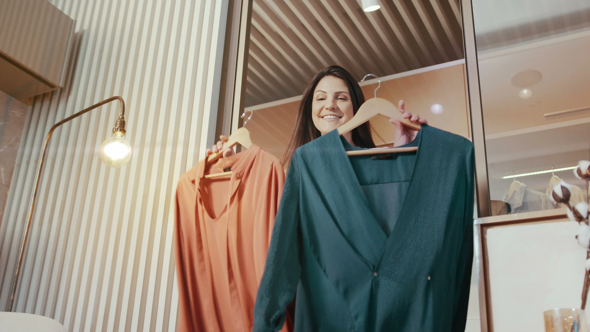 Mulher sorri segurando dois vestidos nos cabides, um laranja e um verde-petróleo, dentro de um closet iluminado.