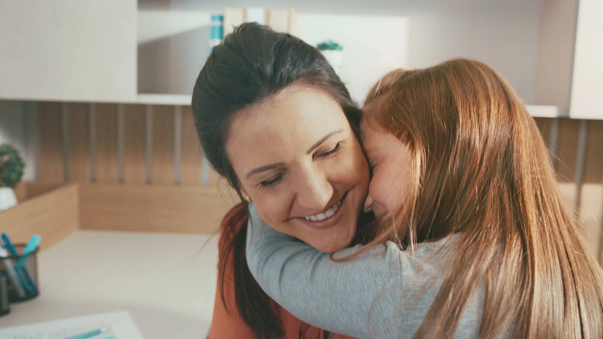 Close de mãe e filha abraçadas, a mãe de olhos fechados e sorriso largo, em ambiente de luz suave.
