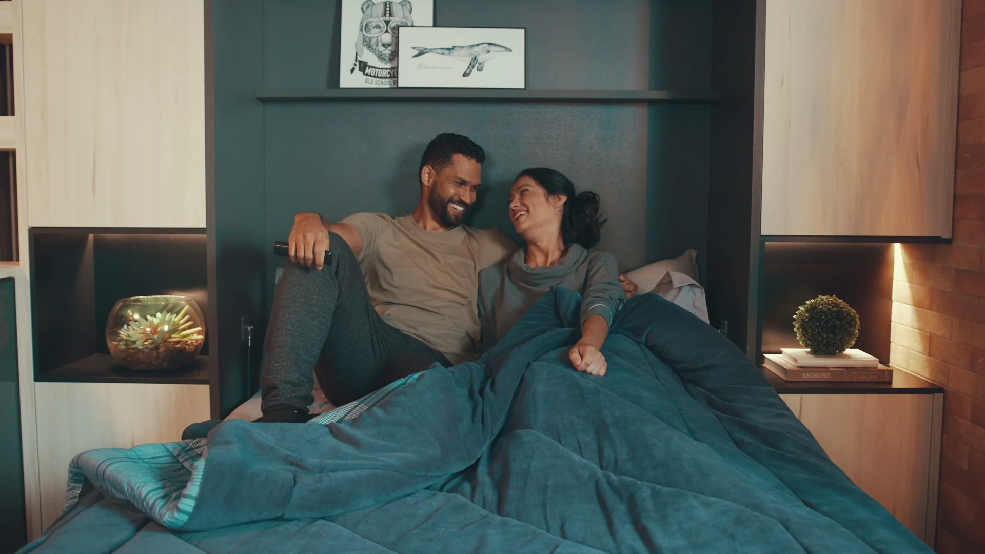 Casal sorri sentado na cama sob um edredom verde-petróleo, em quarto com cabeceira de marcenaria escura e nichos iluminados.