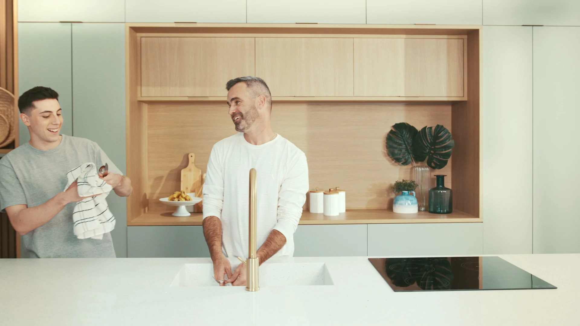 Dois homens riem lavando a louça numa cozinha clara com nicho de madeira, plantas e cooktop preto.