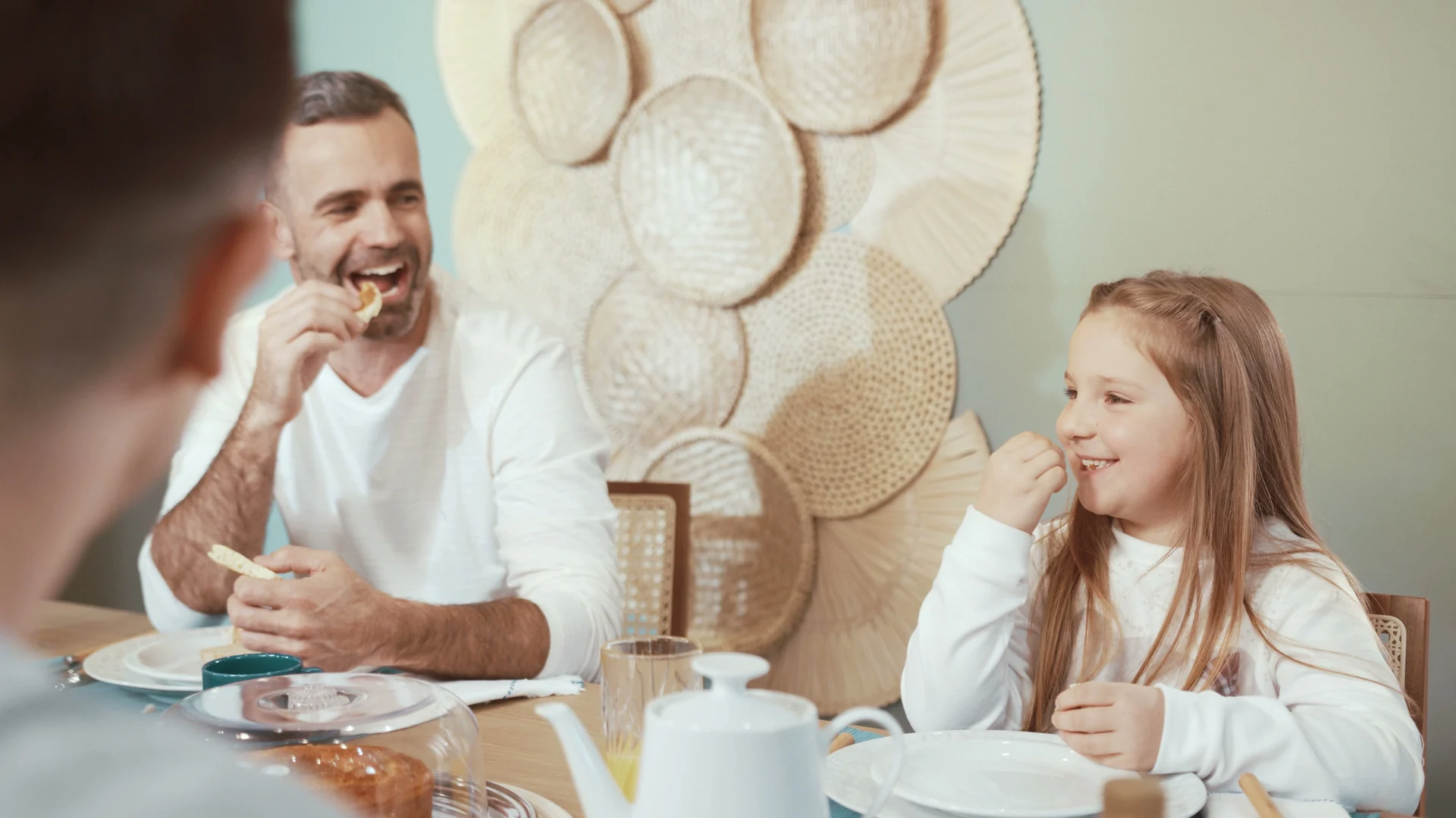 Pai e filha riem à mesa do café da manhã, com bule branco em primeiro plano e painel de cestaria na parede.