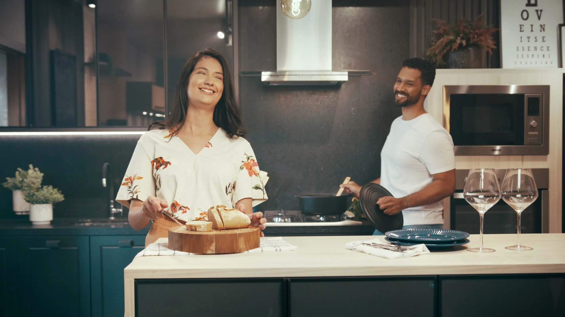 Casal ri na cozinha de armários azul-petróleo: ela serve pão numa tábua na ilha, ele segura uma panela junto ao fogão, com taças e micro-ondas ao fundo.