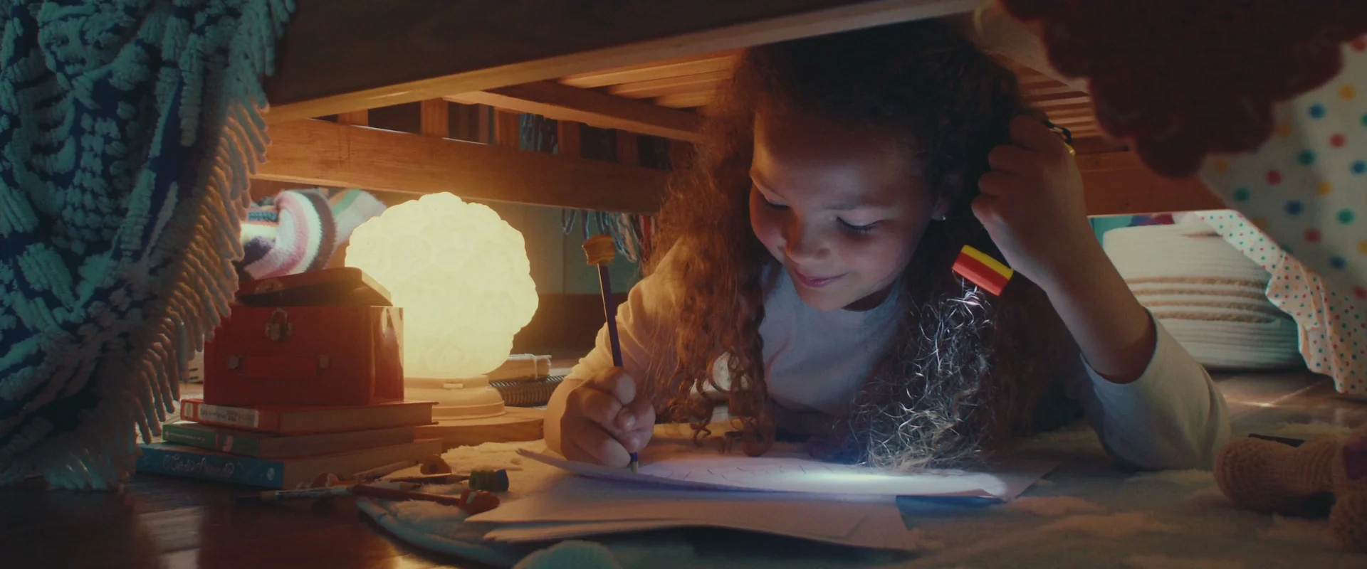 Menina de cabelos cacheados desenha sob uma cama alta, iluminada por uma lanterna e um abajur.