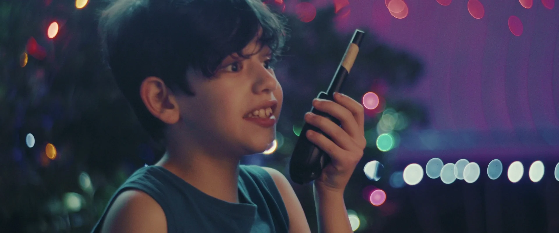 Menino de regata azul fala num walkie-talkie diante de árvore de Natal com luzes coloridas.