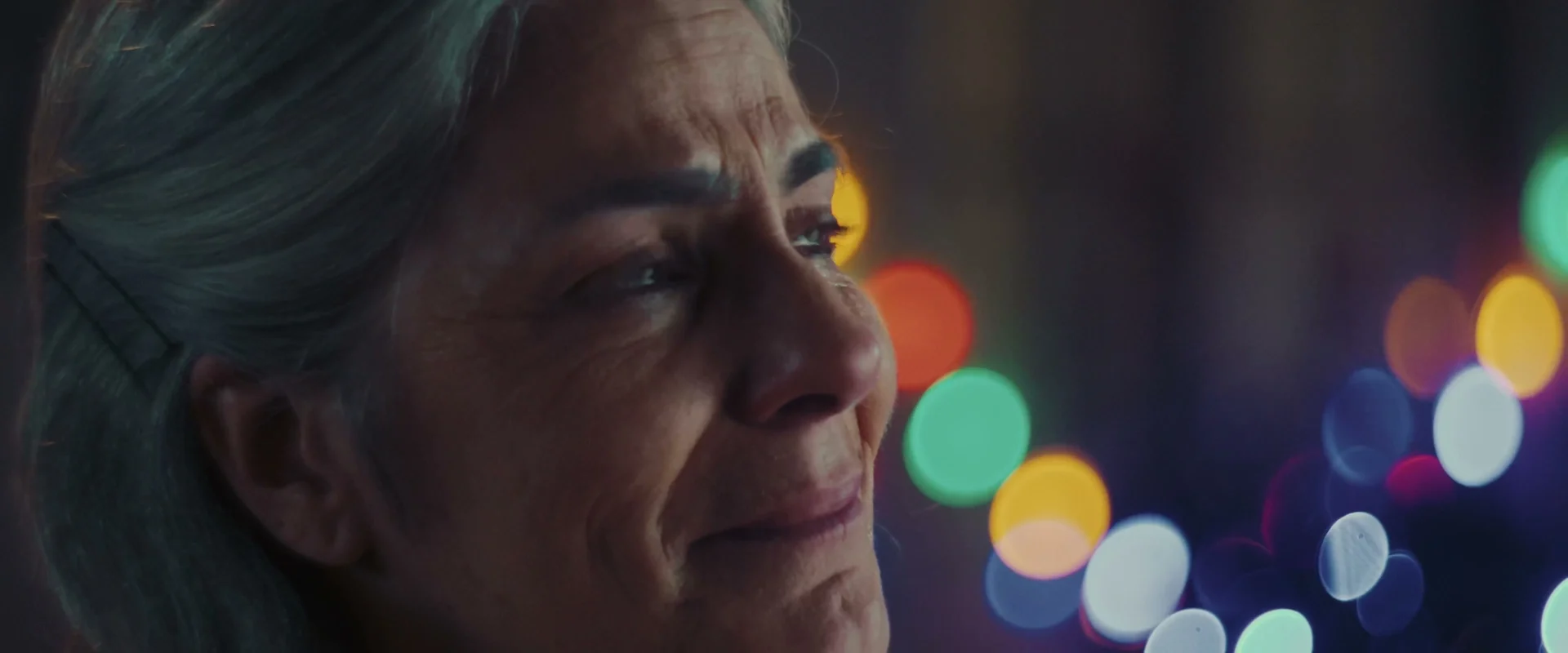 Close da senhora de cabelos grisalhos, emocionada, com luzes de Natal desfocadas em verde, amarelo e azul ao fundo.