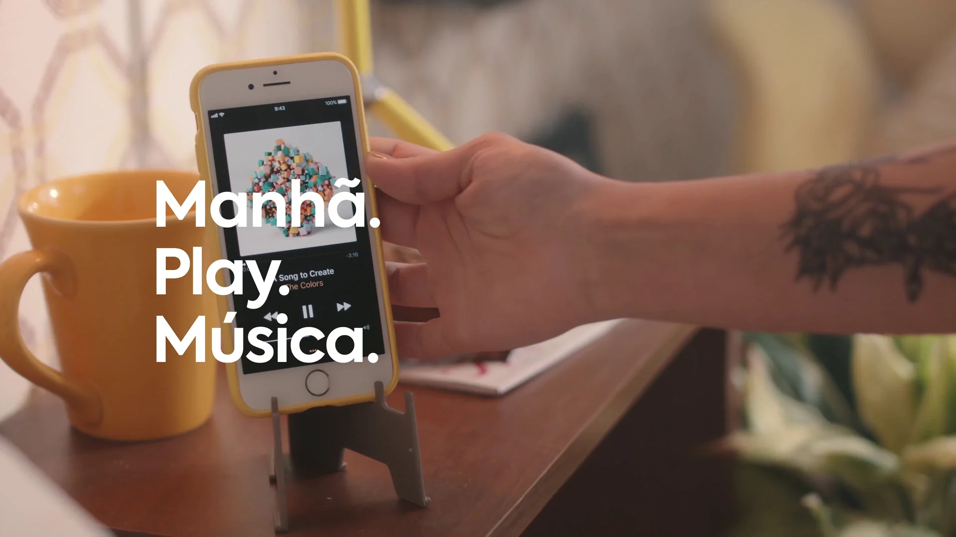 Mão tatuada apoia um celular tocando música em um suporte ao lado de uma caneca amarela, com a cartela Manhã. Play. Música.