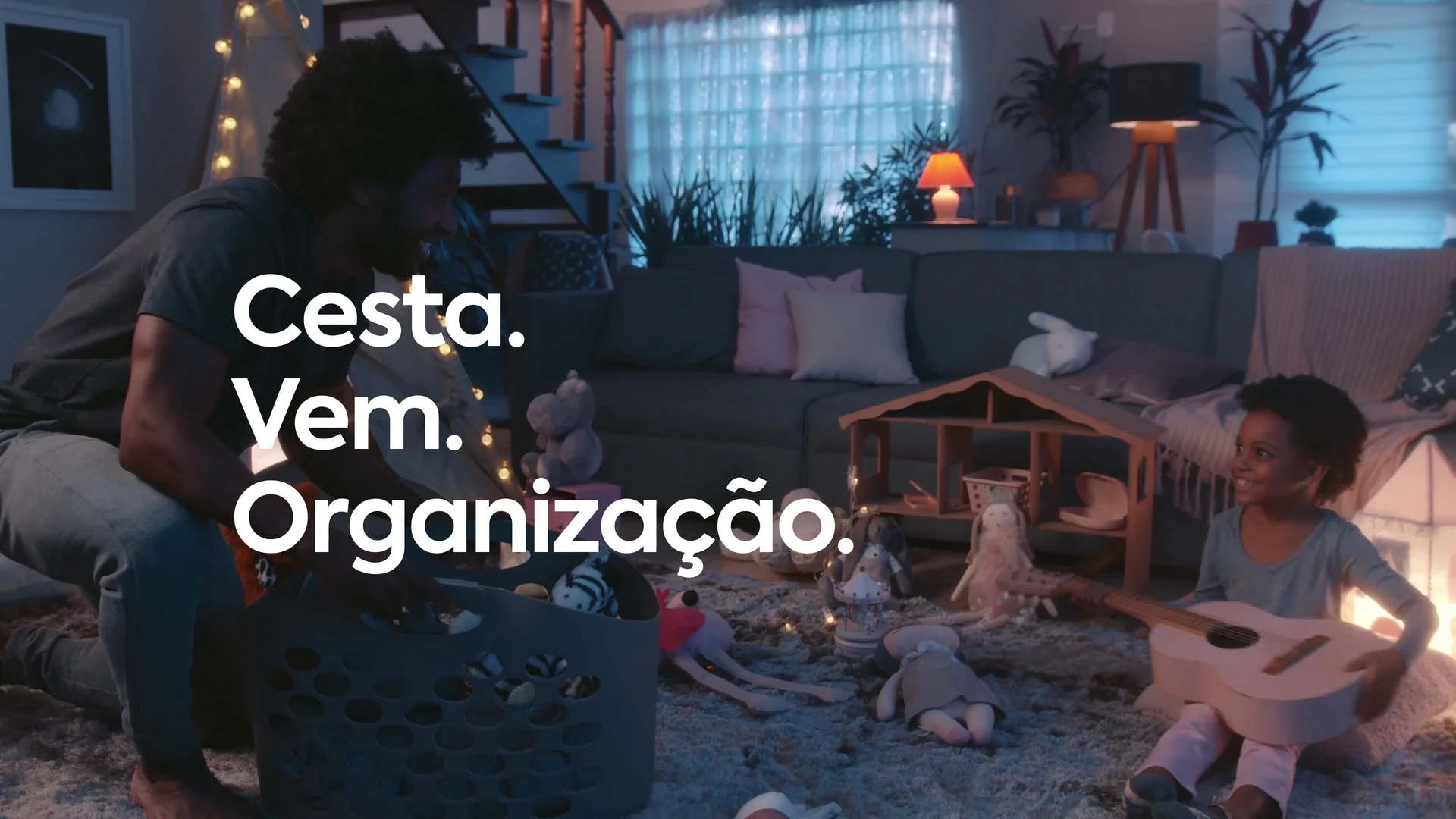 Pai guarda brinquedos em uma cesta enquanto a filha sorri sentada no tapete da sala, com a cartela Cesta. Vem. Organização.
