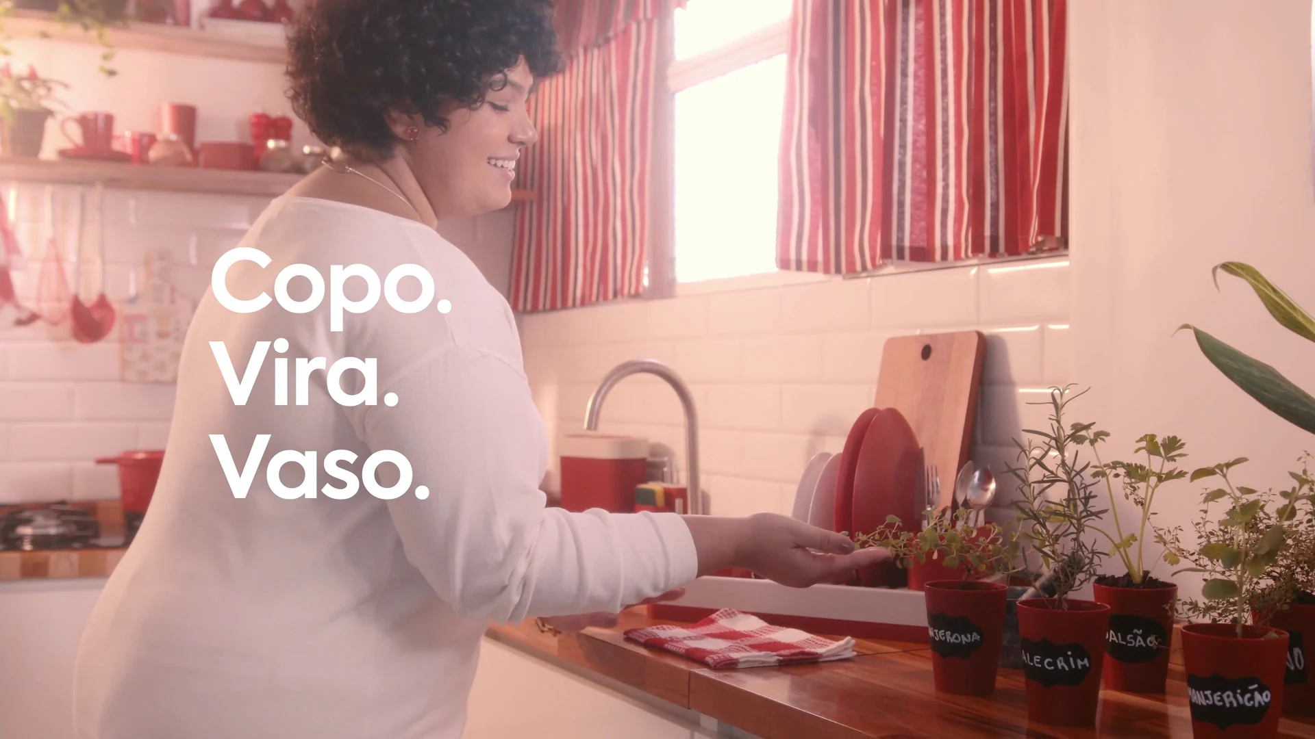 Mulher sorri enquanto ajeita copos-vaso de temperos na bancada de uma cozinha com cortina listrada vermelha.
