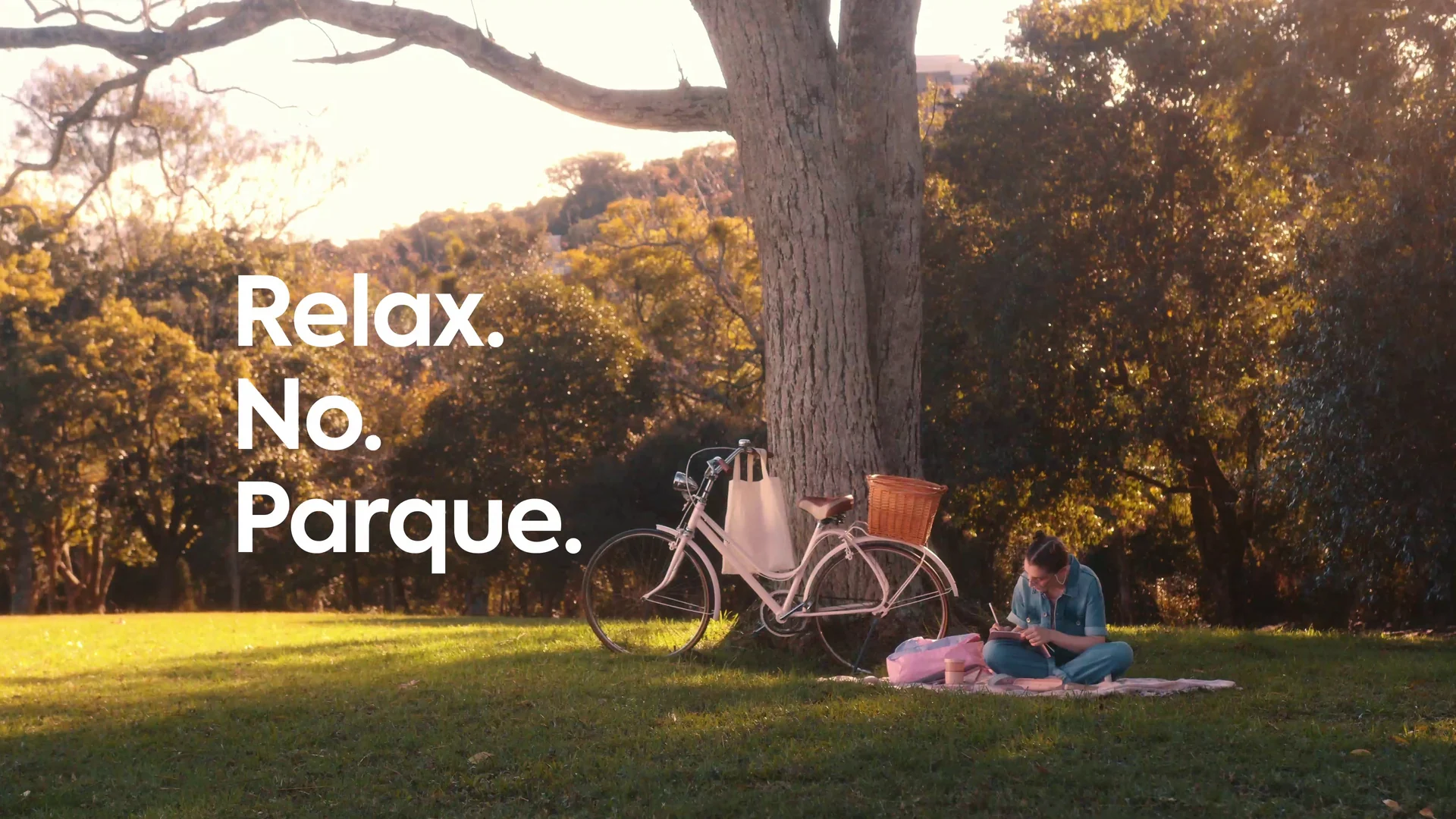 Jovem sentada na grama sob uma árvore, com bicicleta de cestinha ao lado e luz de fim de tarde, com a cartela Relax. No. Parque.