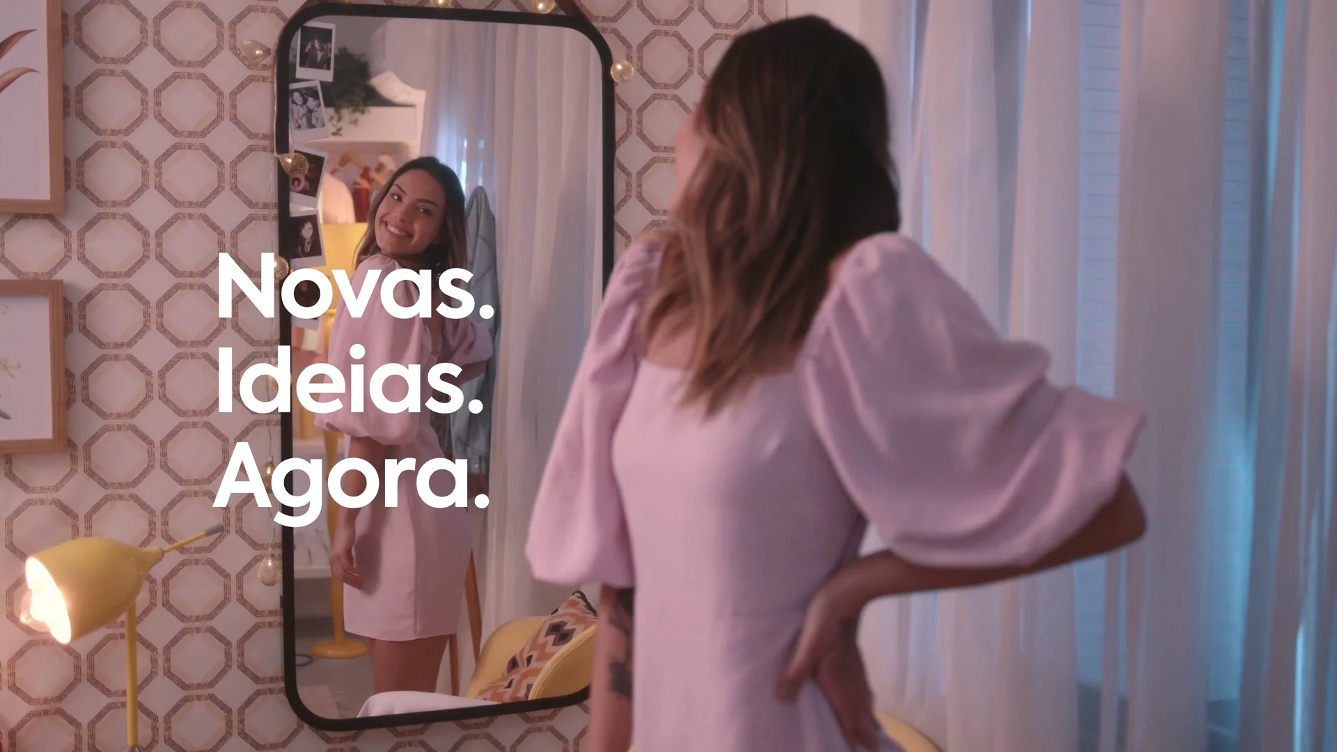 Mulher de blusa rosa se vê num espelho com luzinhas no quarto, com a cartela Novas. Ideias. Agora.