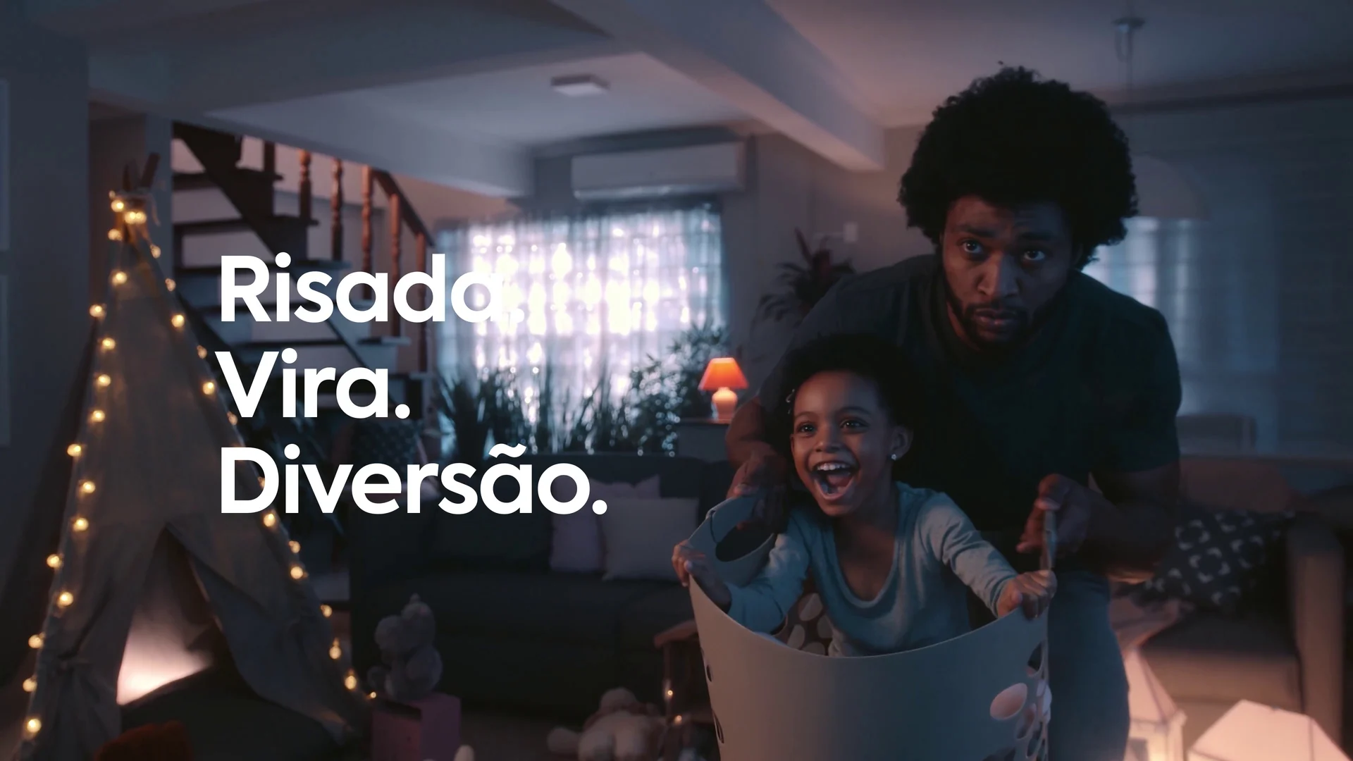 Pai empurra a filha, sorridente dentro de uma cesta, na sala iluminada por luzinhas, com a cartela Risada. Vira. Diversão.