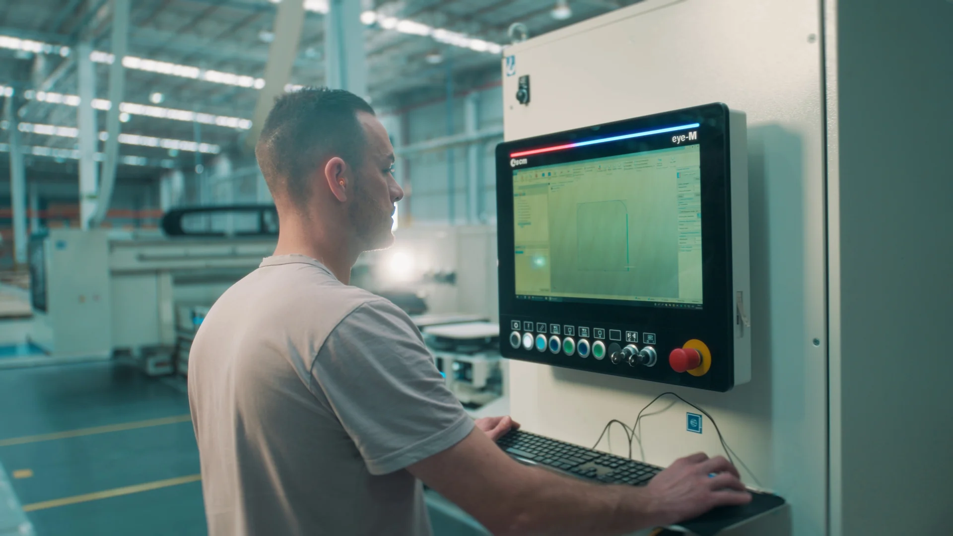 Operador de costas acompanha o painel de controle de uma máquina de usinagem em um galpão iluminado.