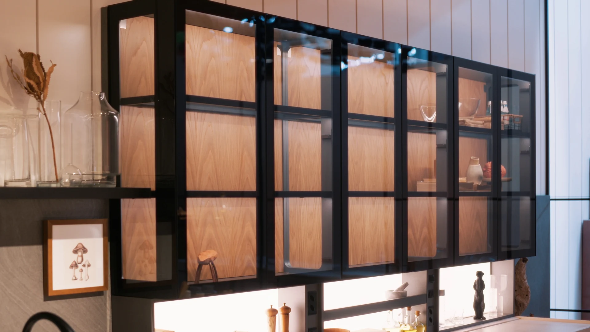 Armário de portas de vidro com estrutura preta e interior de madeira iluminado, com objetos decorativos nas prateleiras.