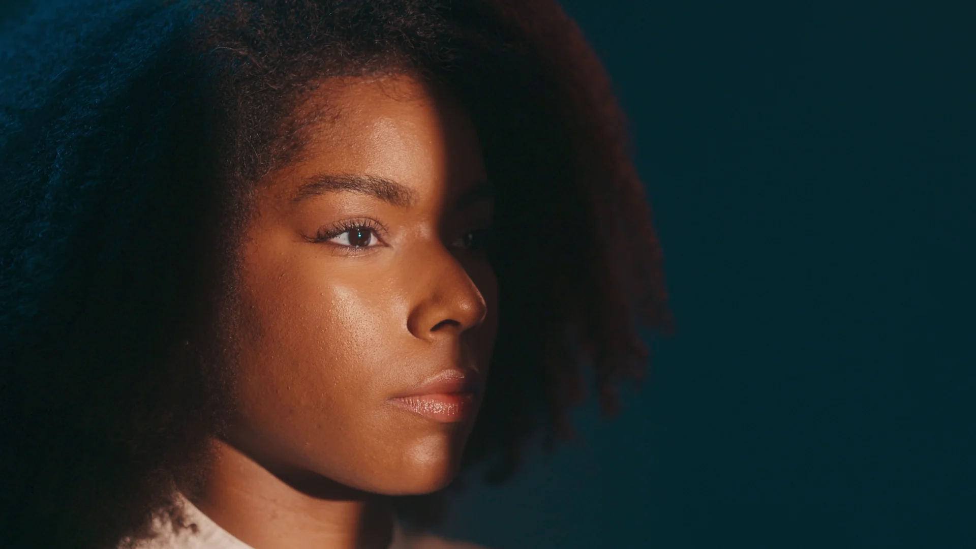 Retrato de perfil de uma mulher de cabelo crespo volumoso, com luz quente no rosto e fundo azul-escuro.