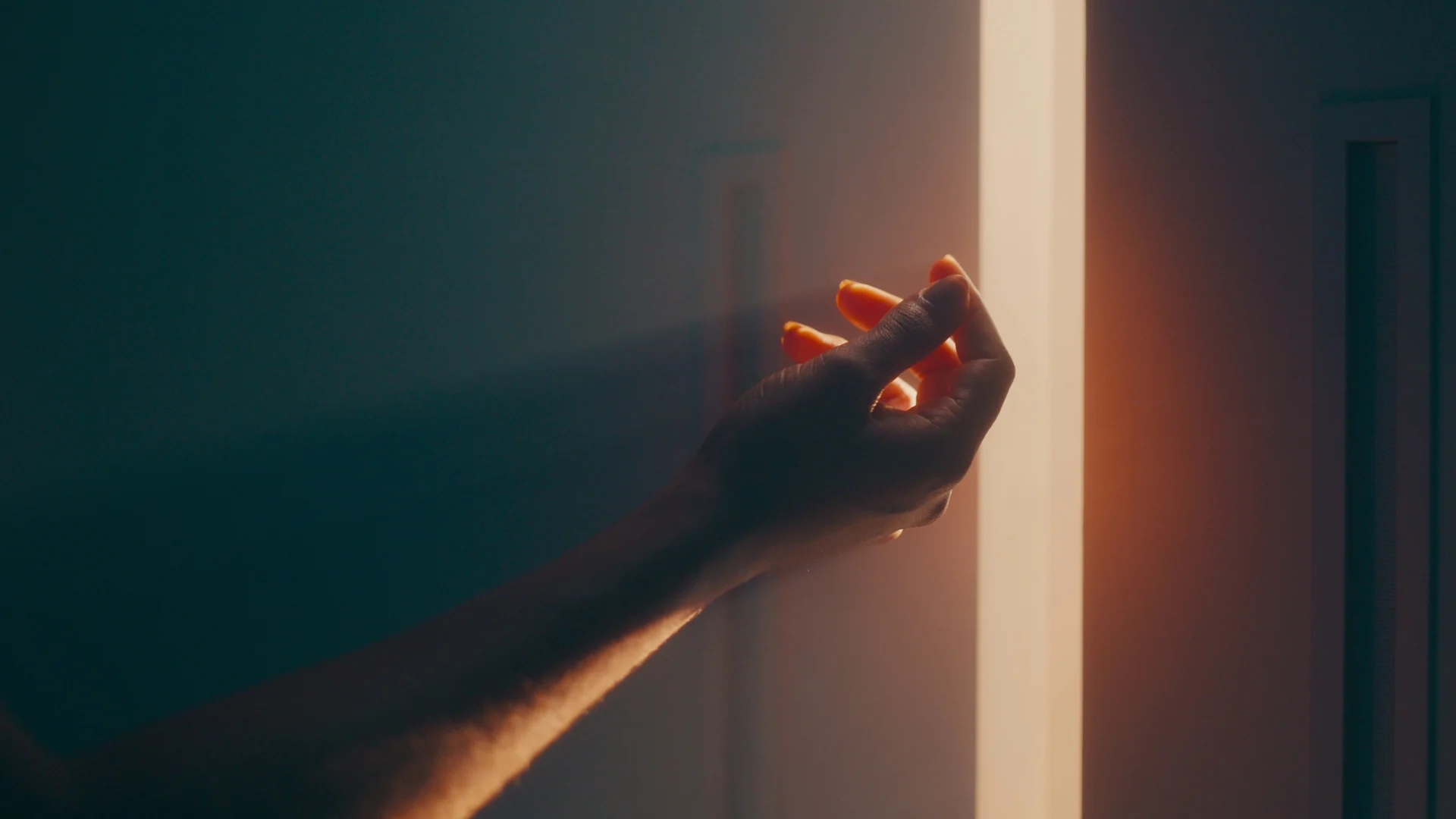 Mão estendida toca um feixe de luz alaranjada que entra pela fresta de uma porta em ambiente escuro.