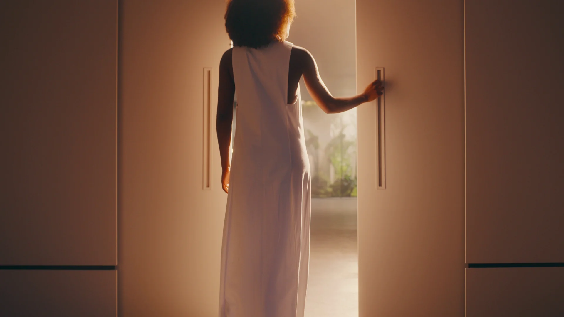Mulher de vestido branco, de costas, abre uma porta de correr e revela um jardim enevoado.