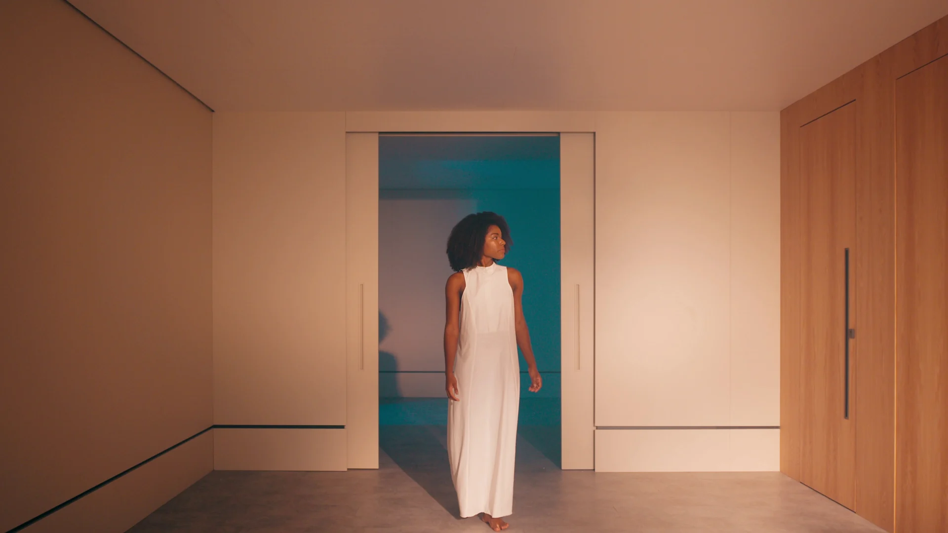 Mulher de vestido branco parada no vão de uma porta embutida, com um cômodo azul atrás e paredes revestidas de madeira.