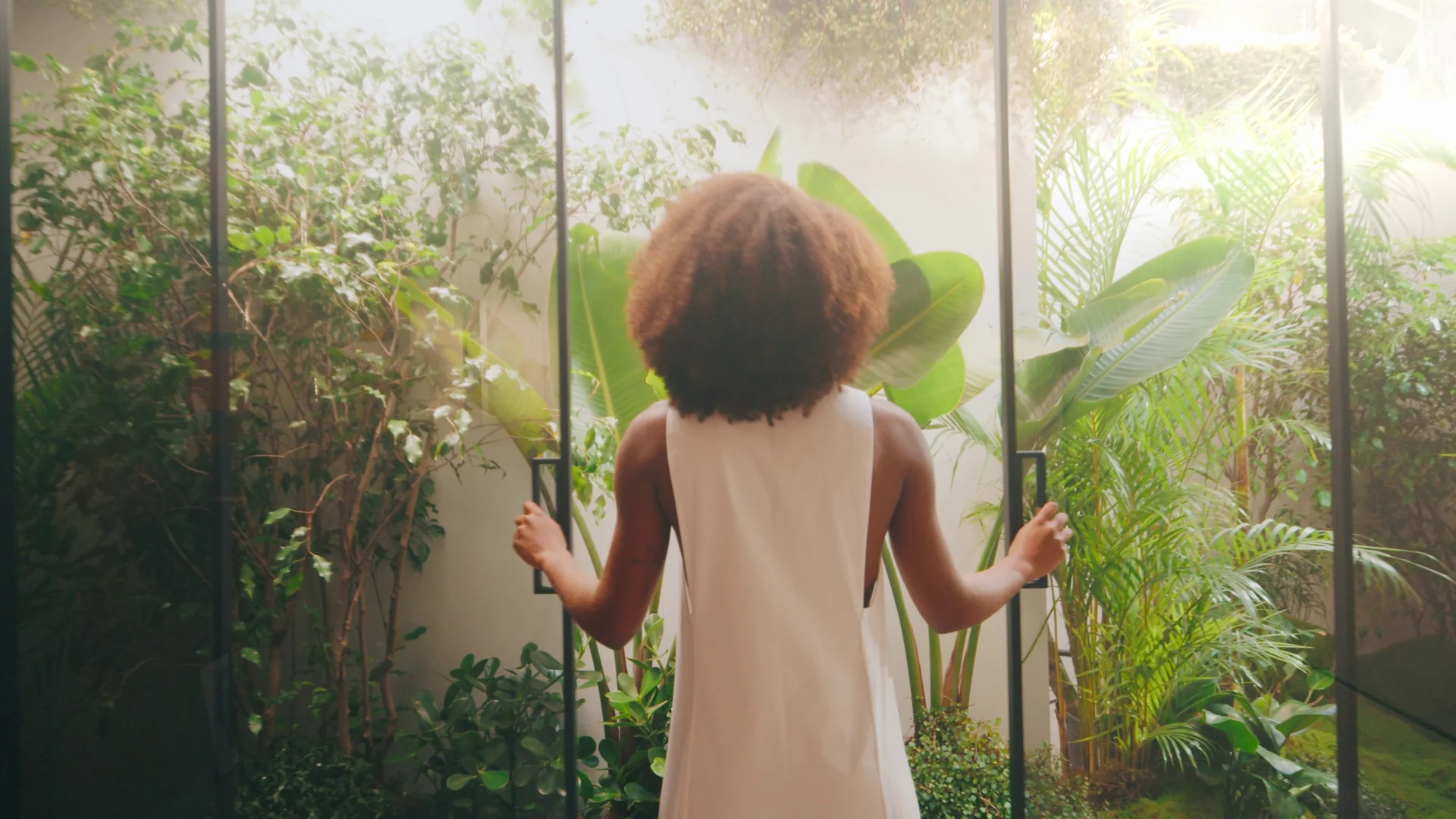 Mulher de costas abre portas de vidro para um jardim interno de folhagens iluminado pelo sol.