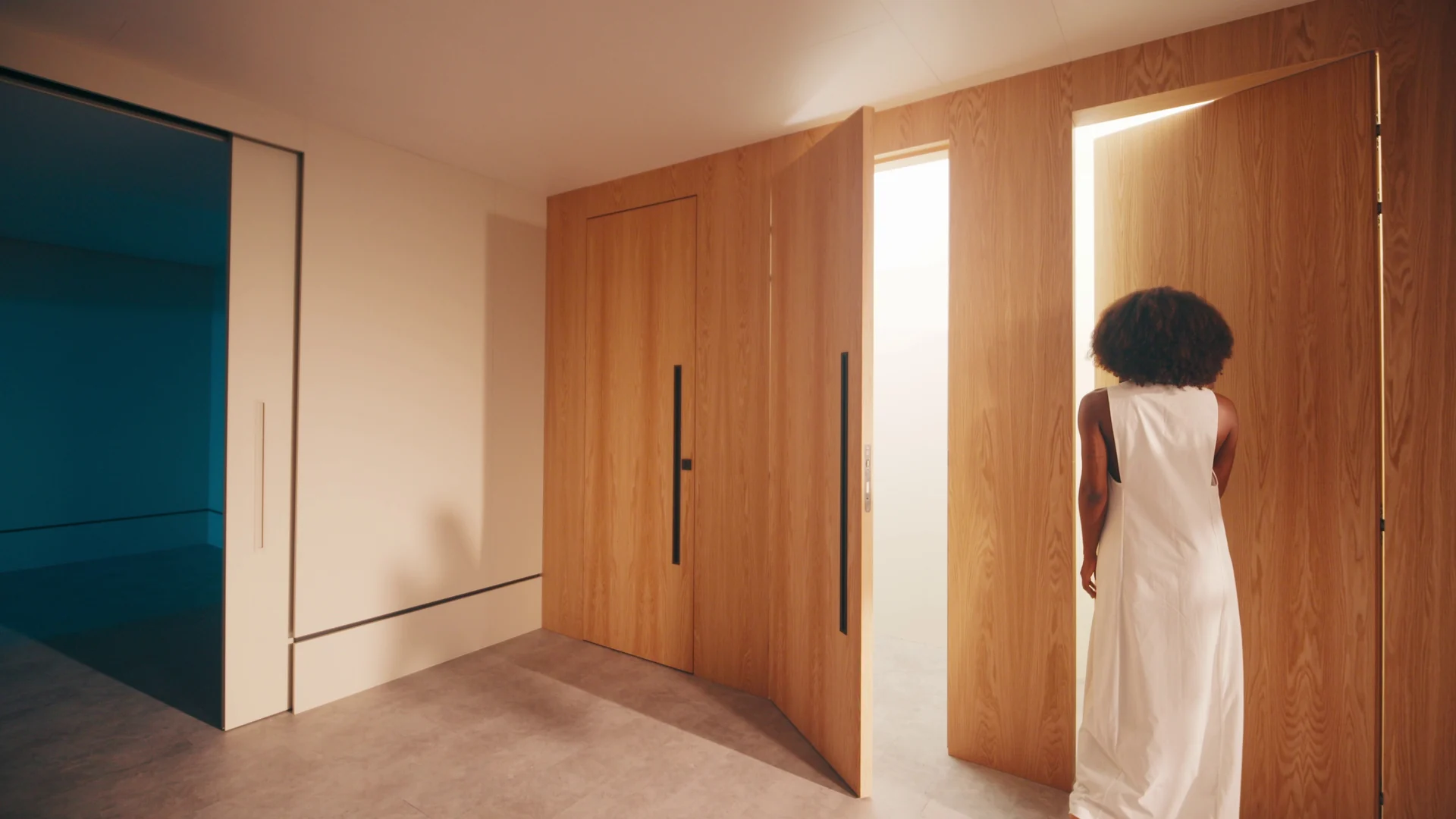 Mulher de vestido branco abre uma grande porta de madeira em um corredor, com um cômodo azulado à esquerda.