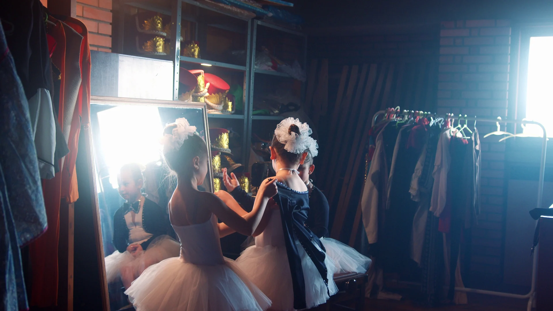 Meninas de tutu e laço no cabelo se arrumam diante de um espelho iluminado em um camarim cheio de figurinos.