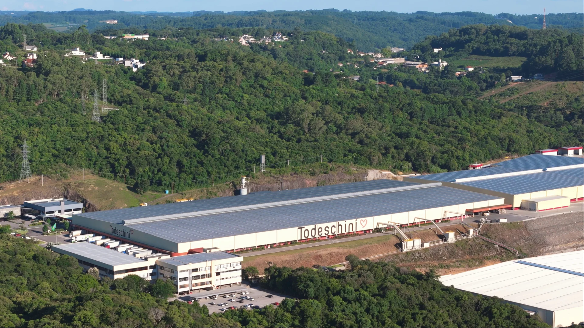 Vista aérea do parque fabril com a fachada Todeschini, cercado por morros cobertos de mata.