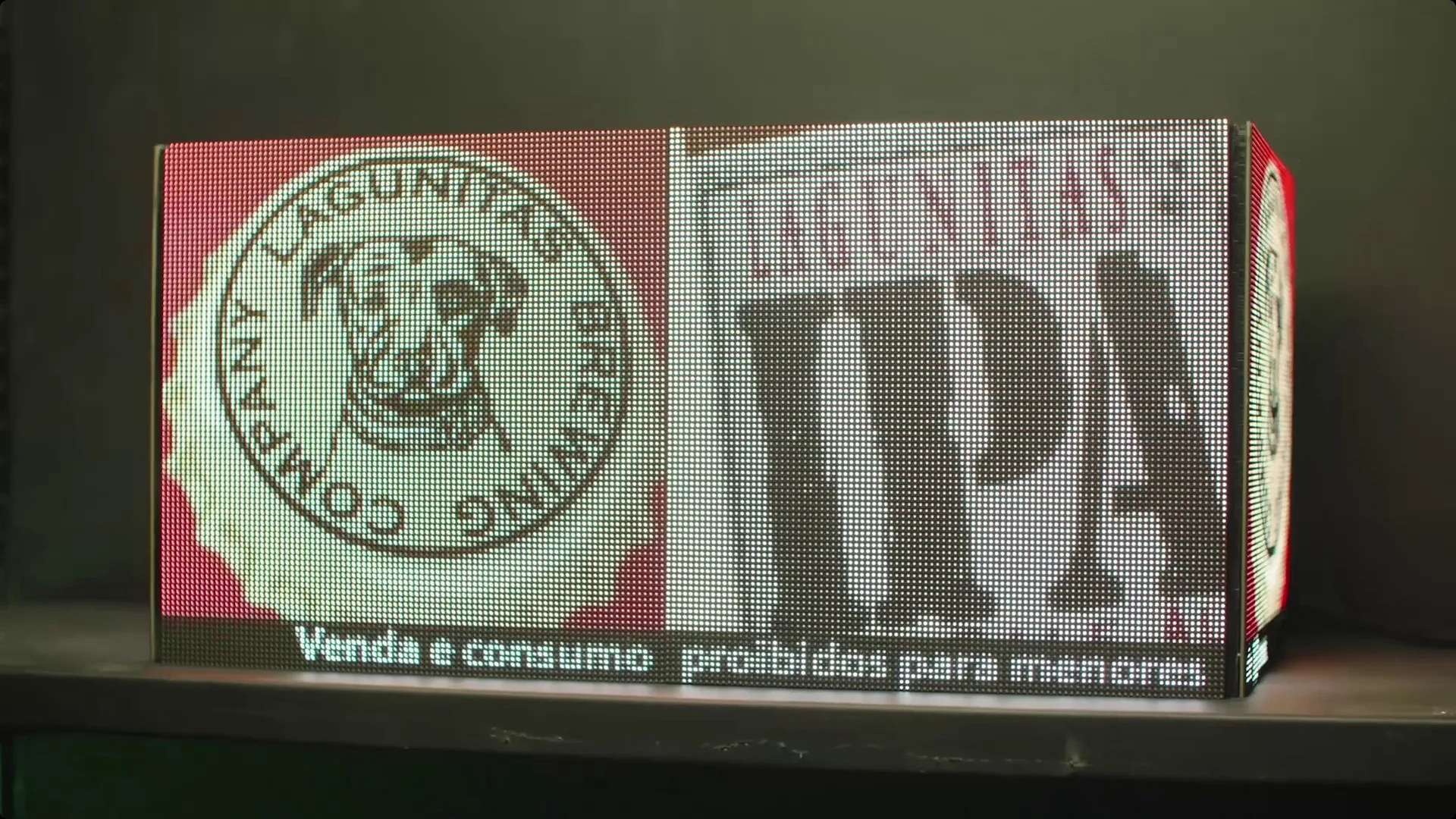 Cubo de telas de LED apoiado sobre uma gôndola exibindo anúncio de cerveja IPA, em fundo verde de estúdio.