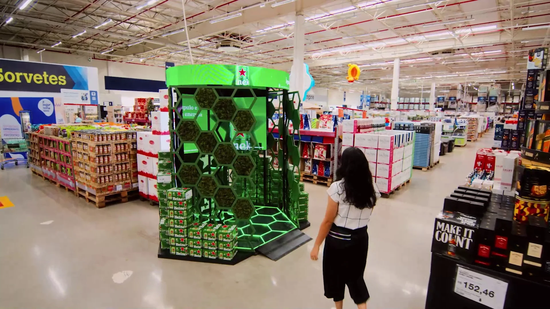 Display hexagonal verde da Heineken com painéis de LED e fardos de cerveja no meio do corredor de um atacarejo, com uma cliente passando ao lado.