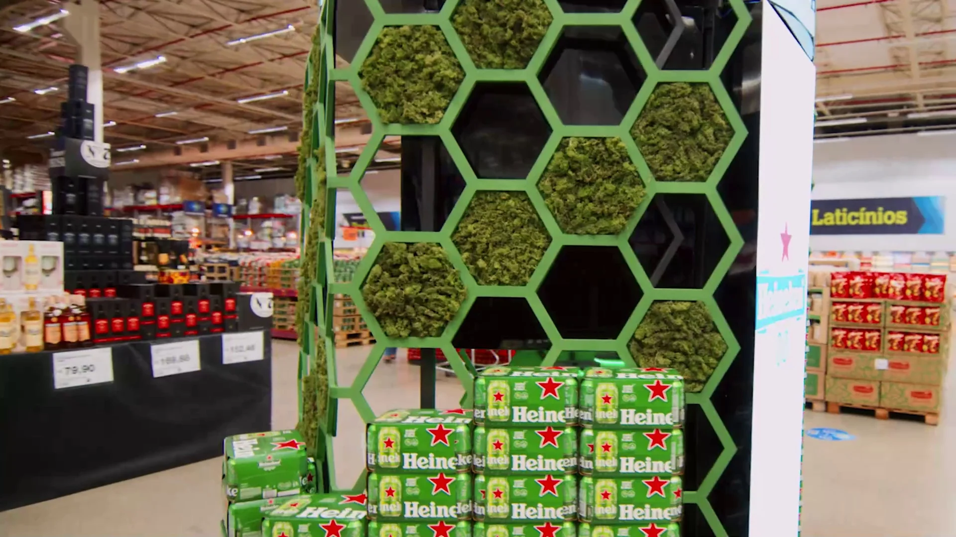 Estrutura hexagonal com musgo e painéis de LED sobre fardos de cerveja Heineken, em corredor de atacarejo.