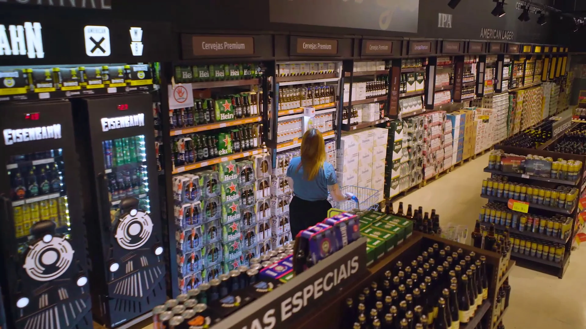Vista alta de um corredor de cervejas especiais em supermercado, com uma consumidora escolhendo produtos junto à gôndola.