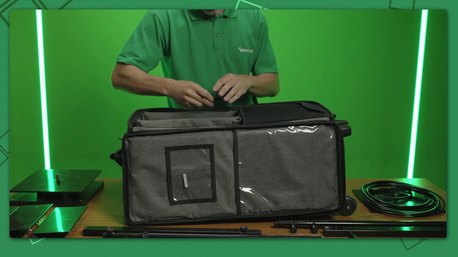 Instrutor de camisa polo verde abre a maleta do kit de telas de LED sobre a bancada, entre bases de chão e hastes, diante de fundo verde de estúdio com tubos de luz.