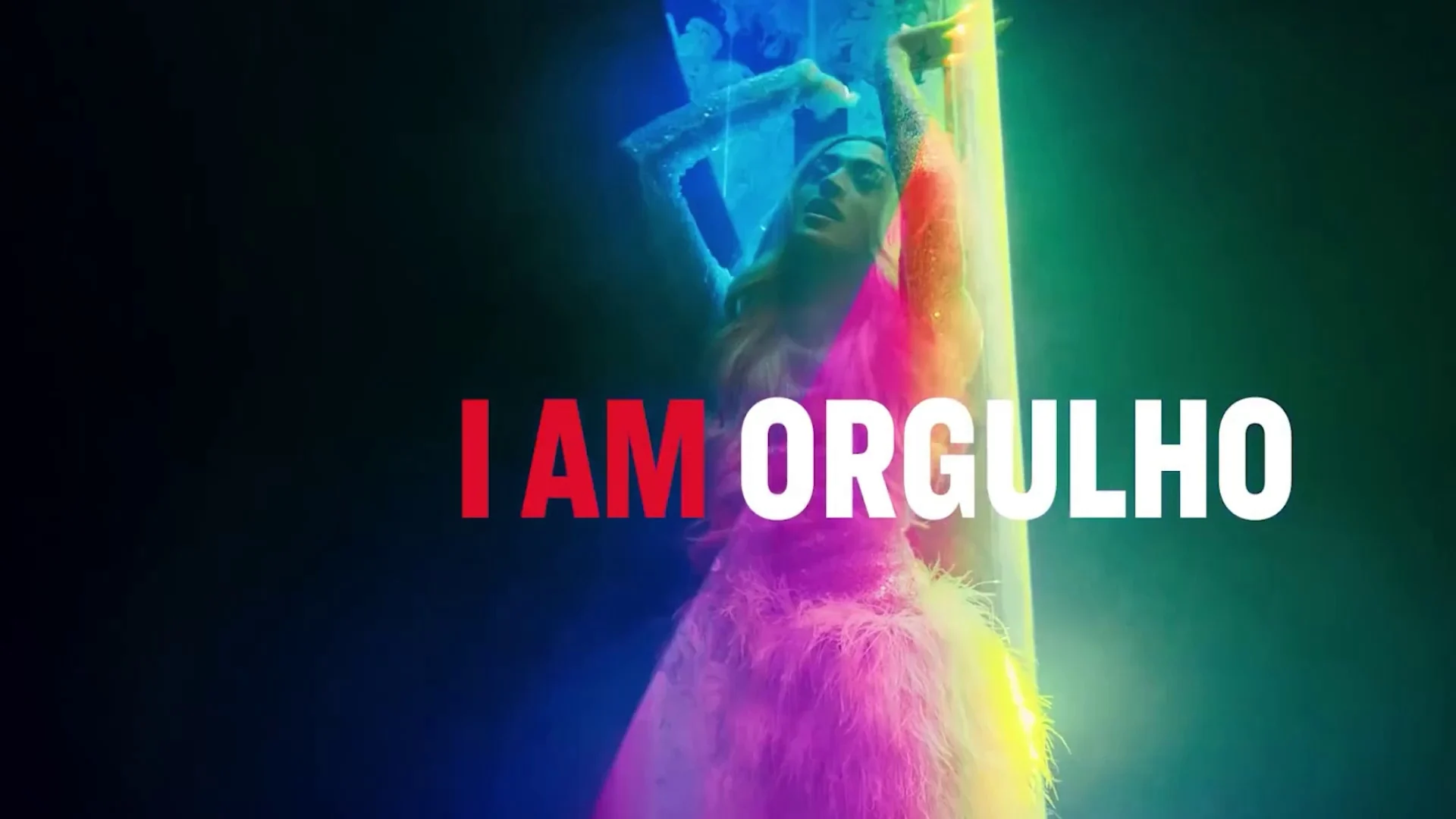 Letreiro "I AM ORGULHO" sobre imagem abstrata em neon colorido, com figura de plumas ao fundo.