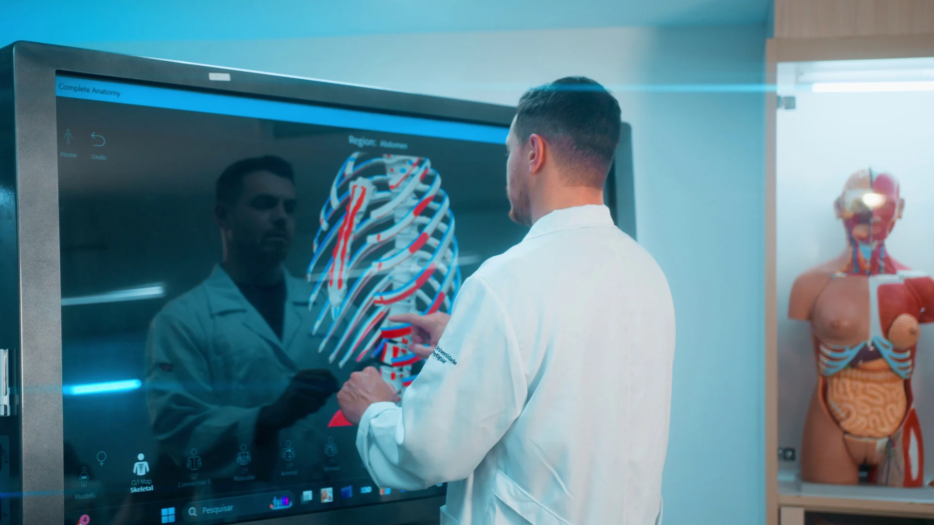 Professor de jaleco aponta a caixa torácica em 3D numa grande tela anatômica, ao lado de um modelo de torso humano.