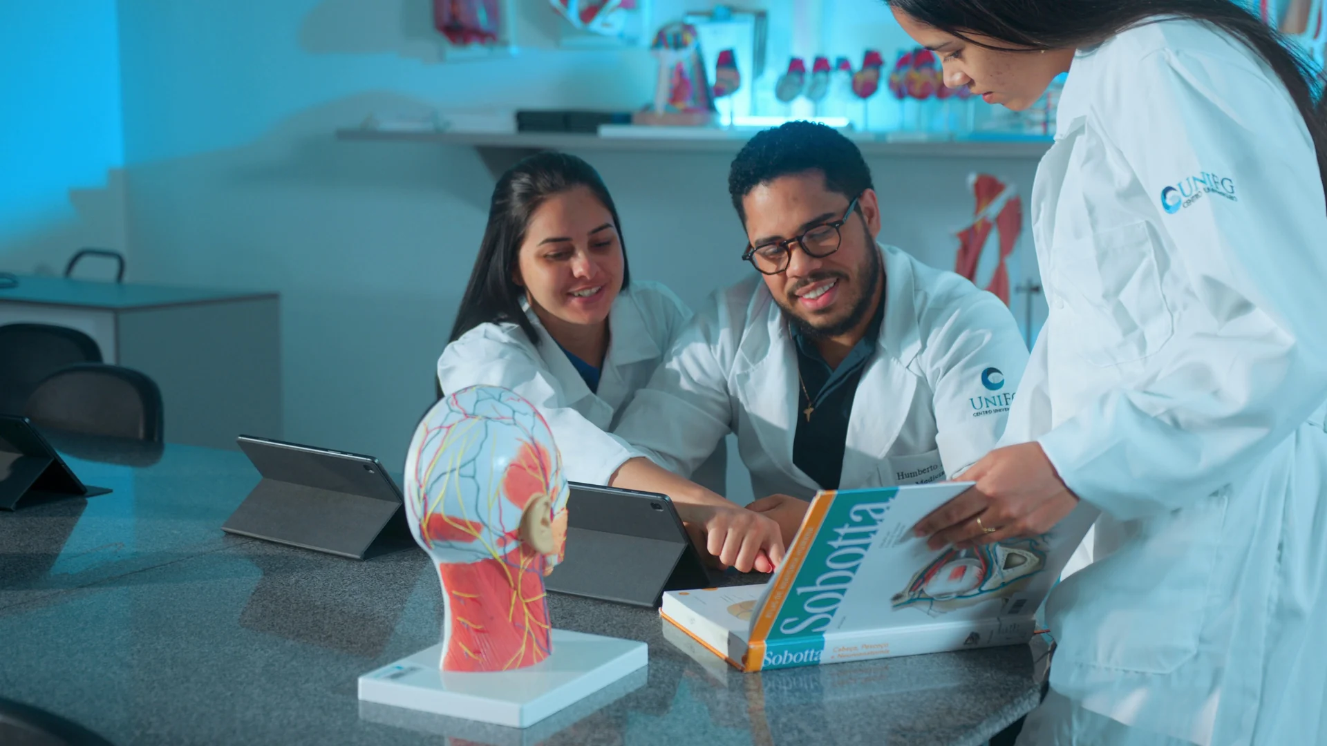 Três estudantes de jaleco sorriem estudando um atlas de anatomia ao lado de um modelo de cabeça humana.