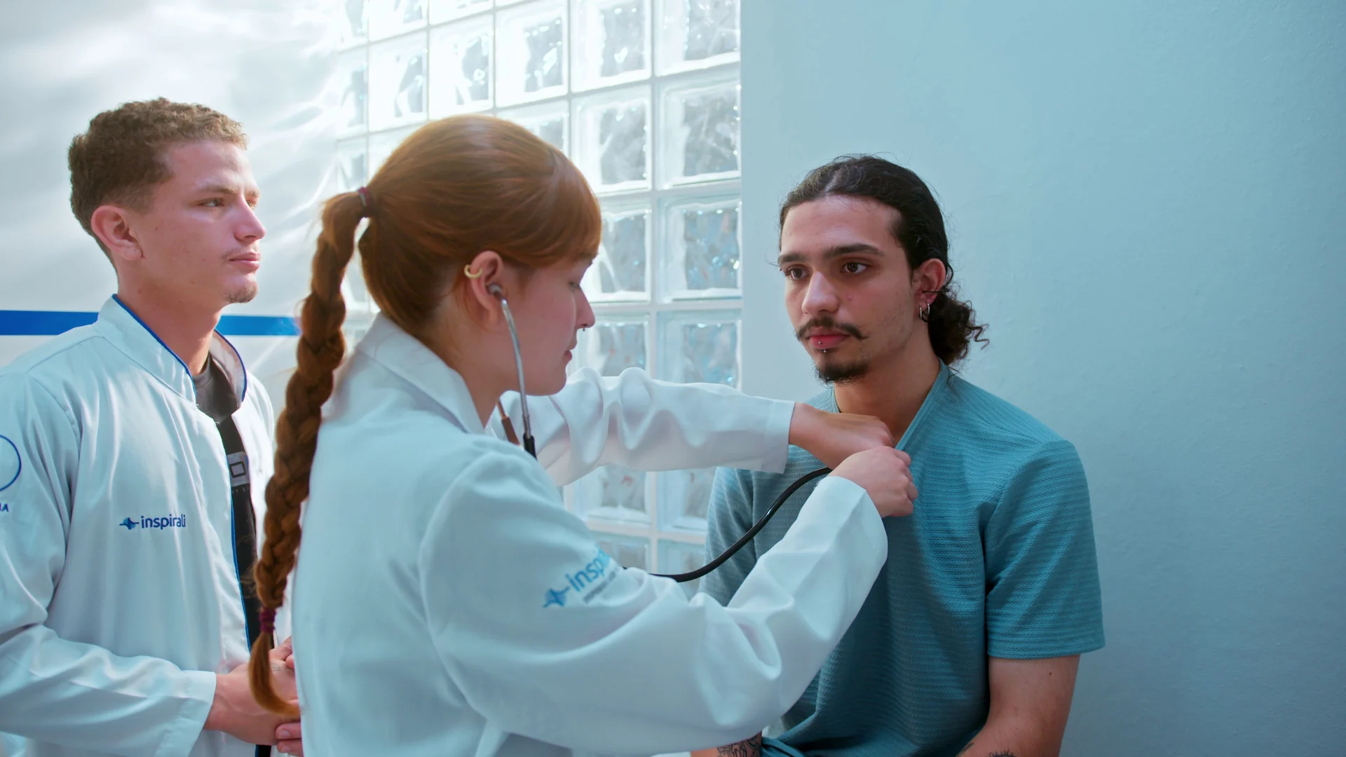 Estudante de trança ausculta um colega com estetoscópio enquanto outro aluno observa.