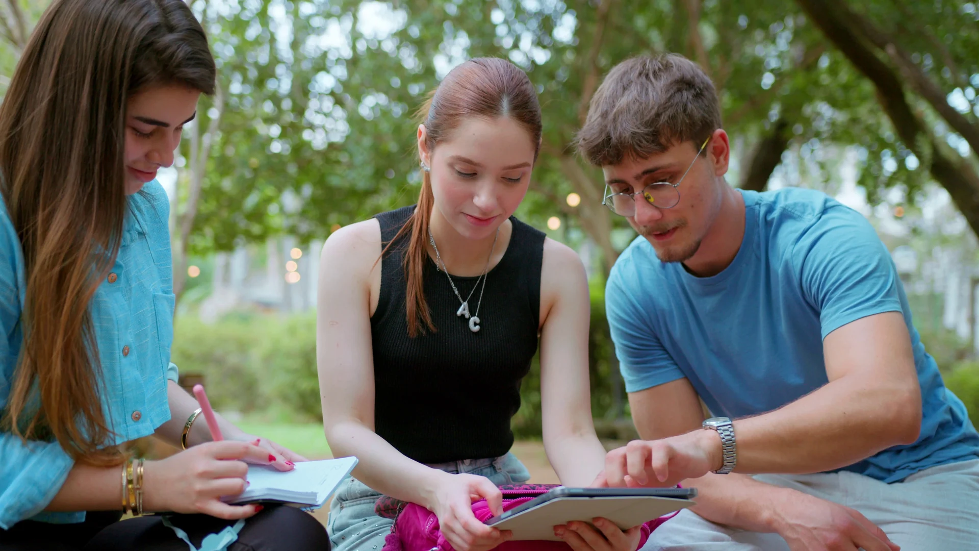 Três estudantes estudam juntos ao ar livre, sob árvores, com caderno e tablet.