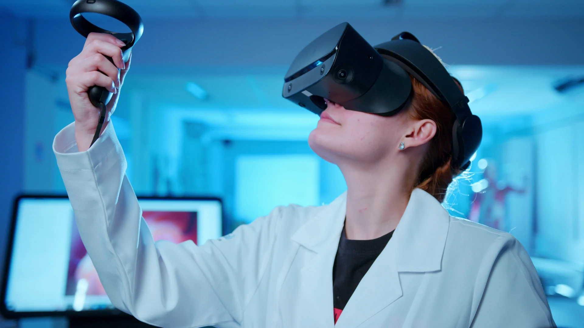 Estudante de jaleco usa óculos de realidade virtual e ergue um controle em um laboratório iluminado de azul.