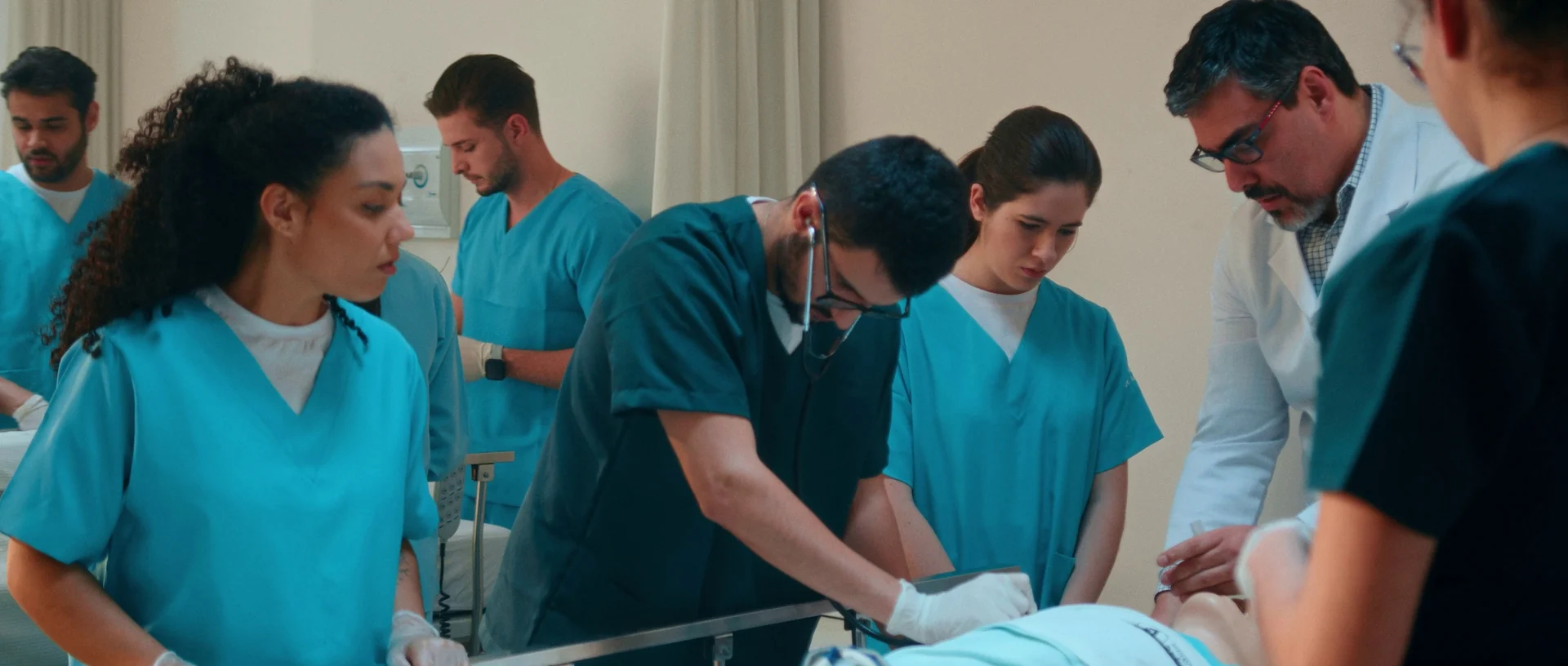 Estudantes de uniforme azul e verde e um professor de jaleco cercam um manequim numa maca durante uma prática de simulação.