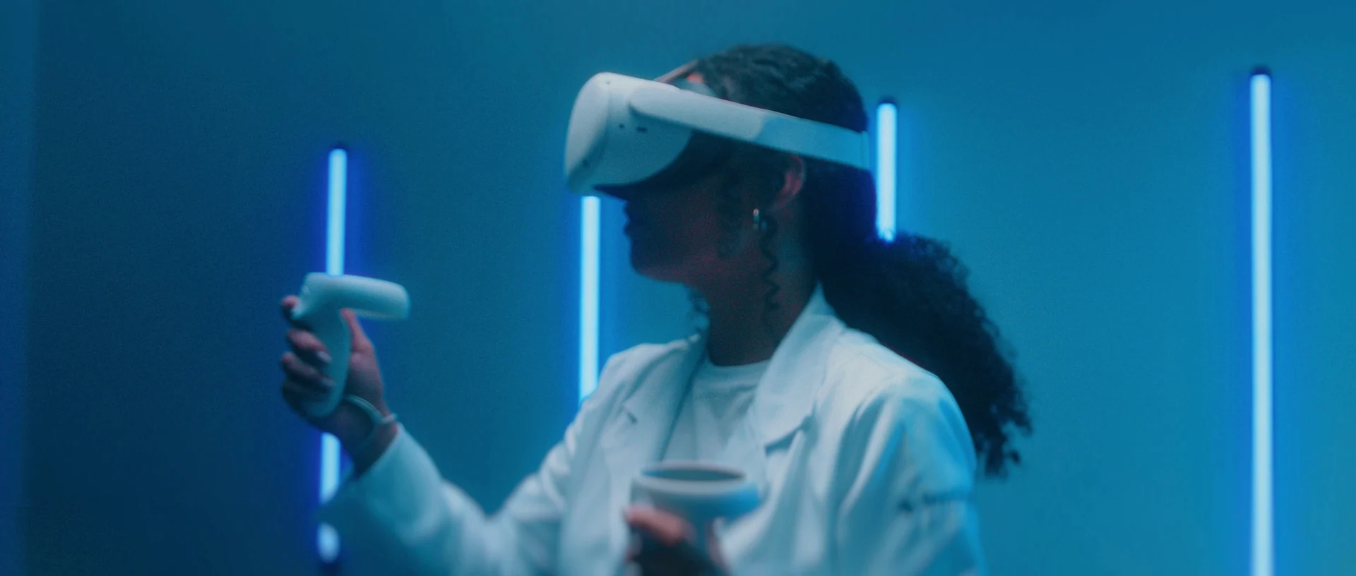 Estudante de jaleco, de perfil, usa óculos de realidade virtual e segura um controle diante de uma parede azul com tiras de luz neon.